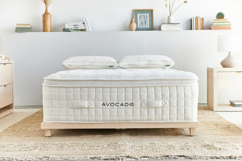 An Avocado mattress shown in a styled bedroom setting, featuring a white quilted mattress cover, two white pillows, and a light wood frame on a woven rug.
